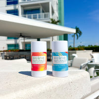 Two Breathe Naturals deodorants, Strawberry Colada and Cooling Cocomint, sit on a ledge near an outdoor pool area under a sunny sky.
