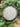 A black bowl contains light gray bath salts, surrounded by fresh mint leaves on a wooden surface.
