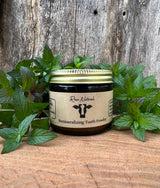 Rowe Naturals presents its remineralizing tooth powder in a glass jar, surrounded by fresh mint leaves, against a rustic wooden background.
