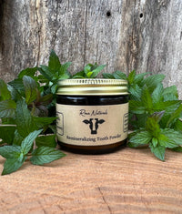 Rowe Naturals presents its remineralizing tooth powder in a glass jar, surrounded by fresh mint leaves, against a rustic wooden background.

