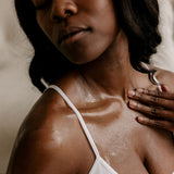 Close-up of a woman with a white strap top, sun-kissed skin, and hand on shoulder.