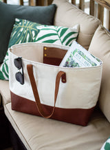 A tan and brown canvas tote bag filled with a book sits on an outdoor patio couch with sunglasses clipped to its side.
