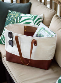 A tan and brown canvas tote bag filled with a book sits on an outdoor patio couch with sunglasses clipped to its side.
