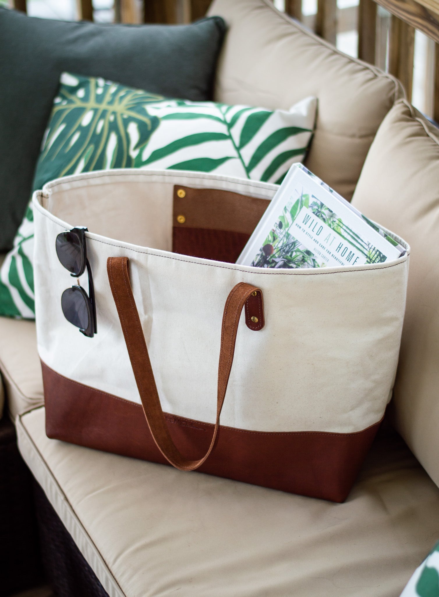 A tan and brown canvas tote bag filled with a book sits on an outdoor patio couch with sunglasses clipped to its side.
