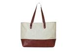 A canvas tote bag with brown leather bottom and straps stands against a white background.
