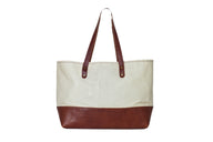 A canvas tote bag with brown leather bottom and straps stands against a white background.
