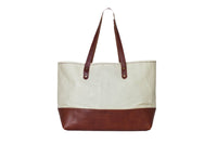 A canvas tote bag with brown leather bottom and straps stands against a white background.
