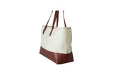 Displays a stylish off-white canvas tote bag featuring a rich brown leather base and handles.
