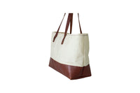Displays a stylish off-white canvas tote bag featuring a rich brown leather base and handles.
