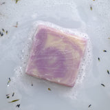 A square of lavender soap rests in bubbly water with scattered lavender buds.
