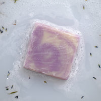 A square of lavender soap rests in bubbly water with scattered lavender buds.
