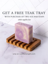A purple swirl bar soap sits on a teak wood soap dish, advertising a free teak tray with a purchase of two 5oz bar soaps from Rustic Strength.
