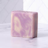 A bar of swirl-patterned lavender and cream artisan soap sits on a marble surface.
