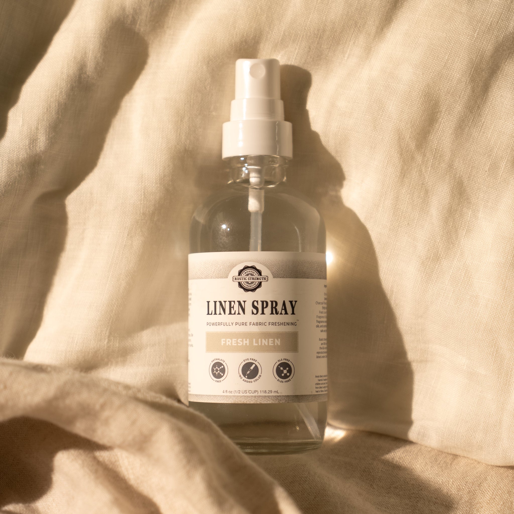 Sunlight illuminates a bottle of Rustic Strength Fresh Linen spray resting on a soft, folded linen fabric.