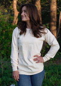 A brunette woman smiles while modeling a cream-colored long-sleeve shirt and jeans outdoors.

