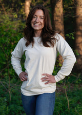 Smiling woman models a long-sleeve cream shirt and blue jeans while standing outdoors in a wooded area.
