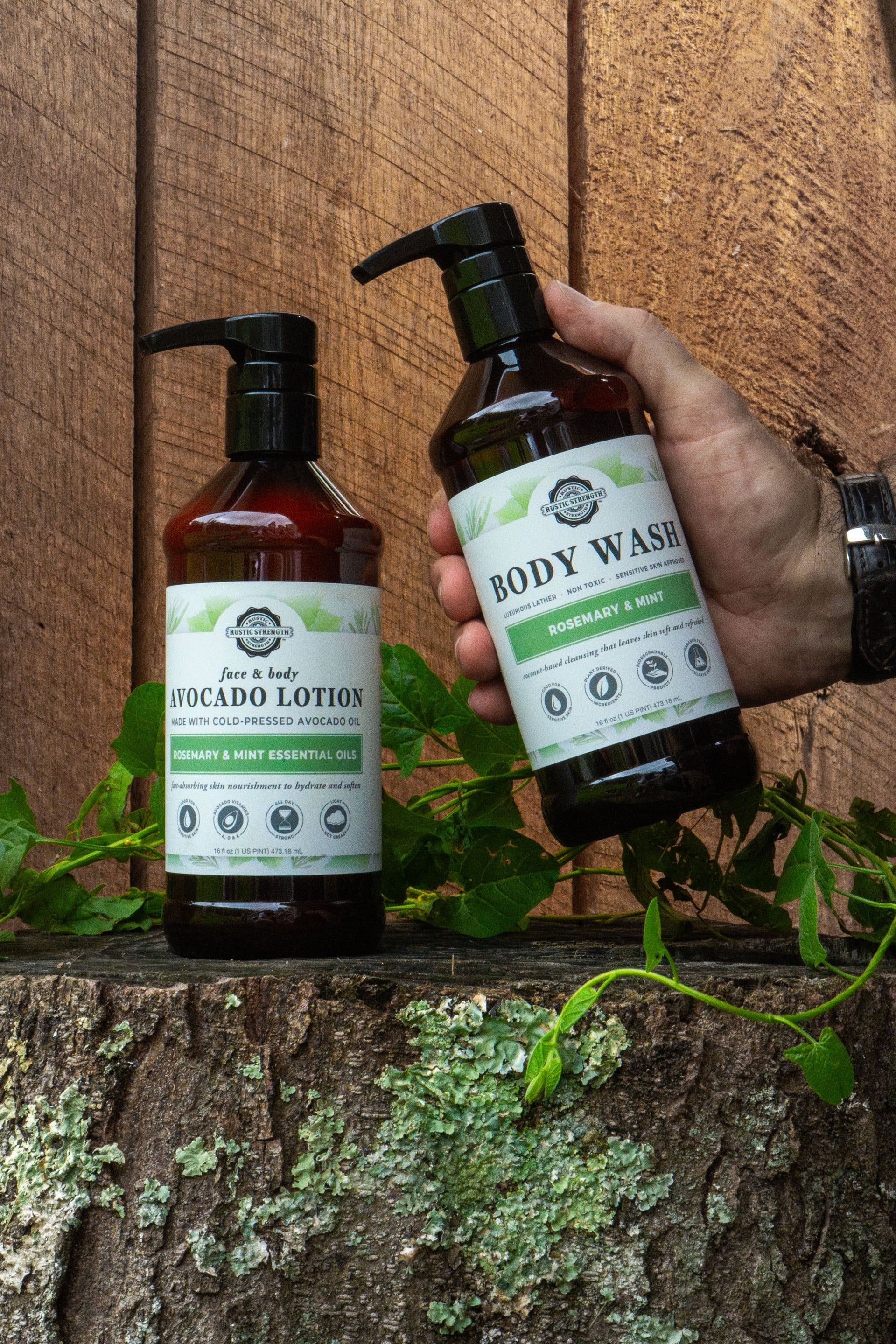 Hand holding a bottle of body wash next to avocado lotion on a wooden surface with greenery.
