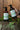 Hand holding a bottle of body wash next to avocado lotion on a wooden surface with greenery.