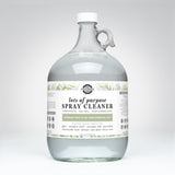 A one-gallon glass jug holds Rustic Strength's Lots of Purpose Spray Cleaner, featuring rosemary mint and tea tree essential oils for a natural cleaning solution.
