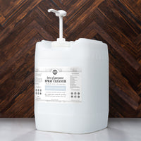 A large container of Rustic Strength all-purpose spray cleaner with pump sits on a marble surface against a dark wood wall.