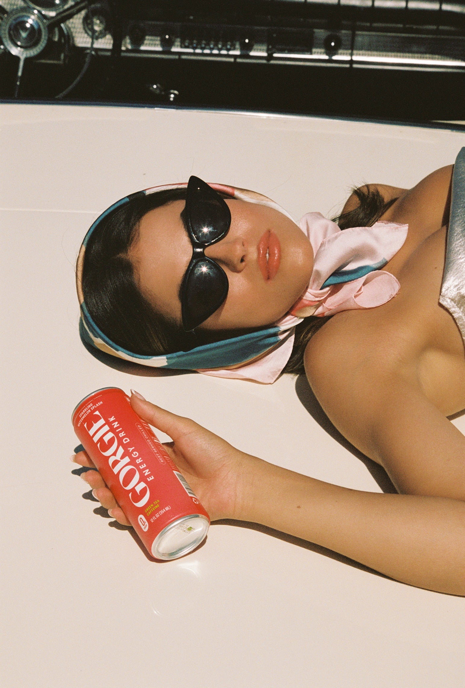 Woman in sunglasses and headscarf reclines on a car, holding a Gorgie energy drink can.
