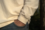 Person stands with hand casually in denim jeans pocket while wearing a cream-colored long-sleeved shirt.
