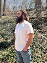 A bearded man wearing a white t-shirt stands outside with hands in his jeans pockets, looking towards trees and a blue sky.

