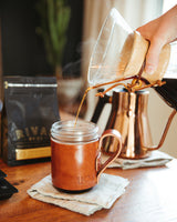 Pouring hot Riva Bros Coffee into a leather-sleeved mug on a wooden table.

