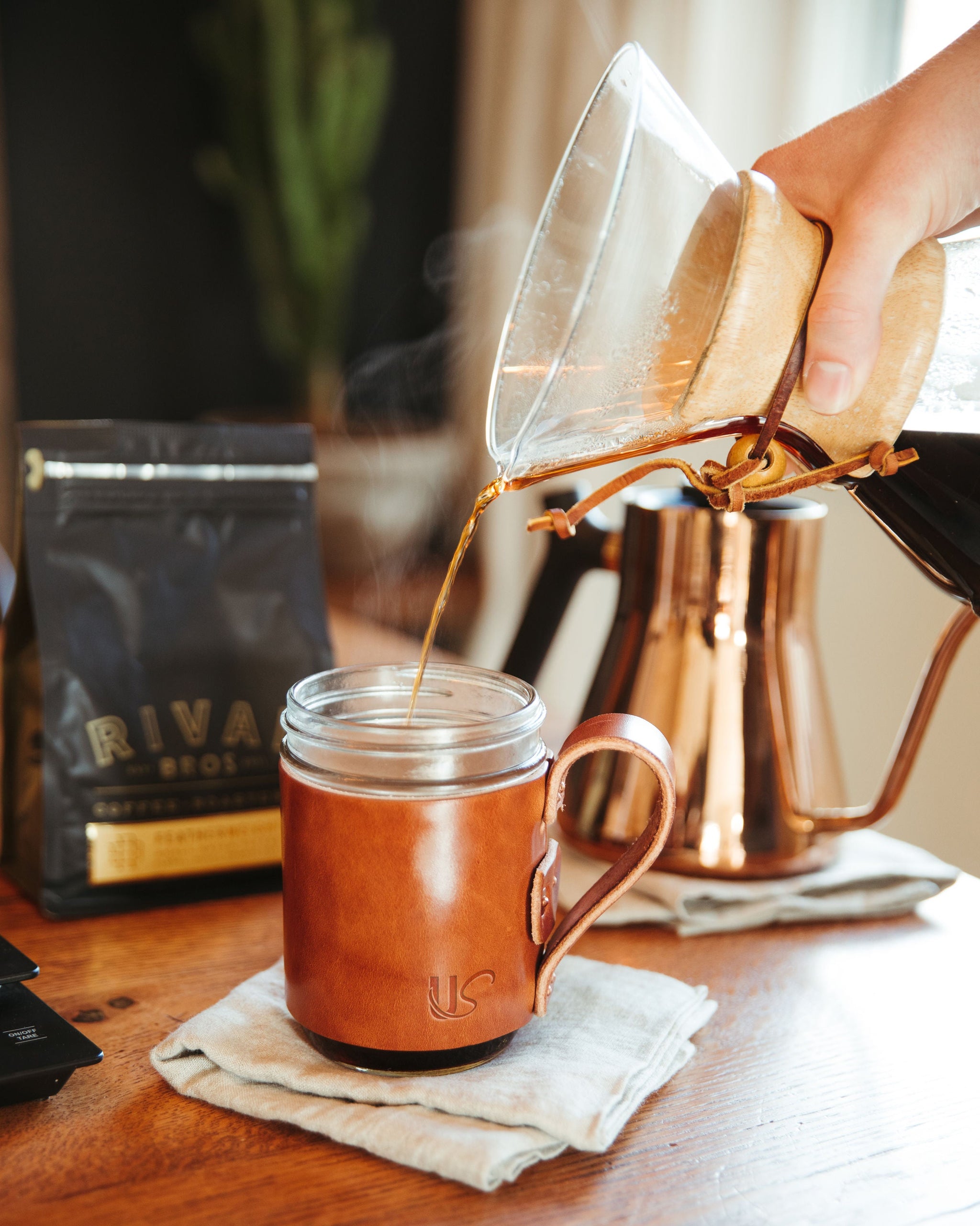 Pouring hot Riva Bros Coffee into a leather-sleeved mug on a wooden table.
