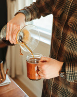 A man pours fresh, dark roast coffee from a glass Chemex carafe into a leather-sleeved mug.
