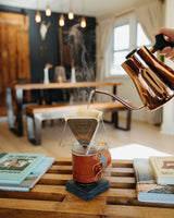 Someone pours hot water from a copper kettle into a pour-over coffee maker on a leather-sleeved mug, steaming in a cozy home setting.
