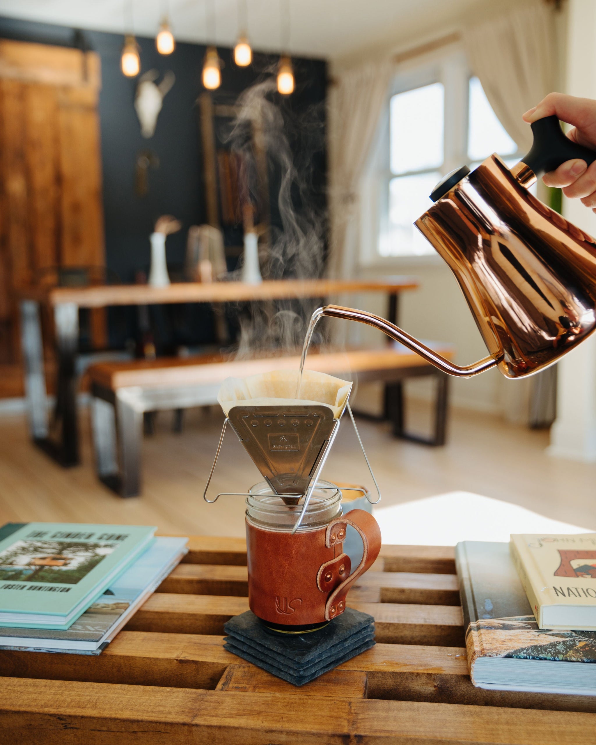 Someone pours hot water from a copper kettle into a pour-over coffee maker on a leather-sleeved mug, steaming in a cozy home setting.

