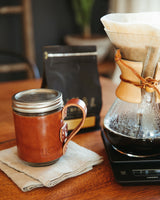 Pour-over coffee brews into a glass carafe on a scale beside a leather-sleeved mason jar coffee mug and a bag of roasted coffee beans.