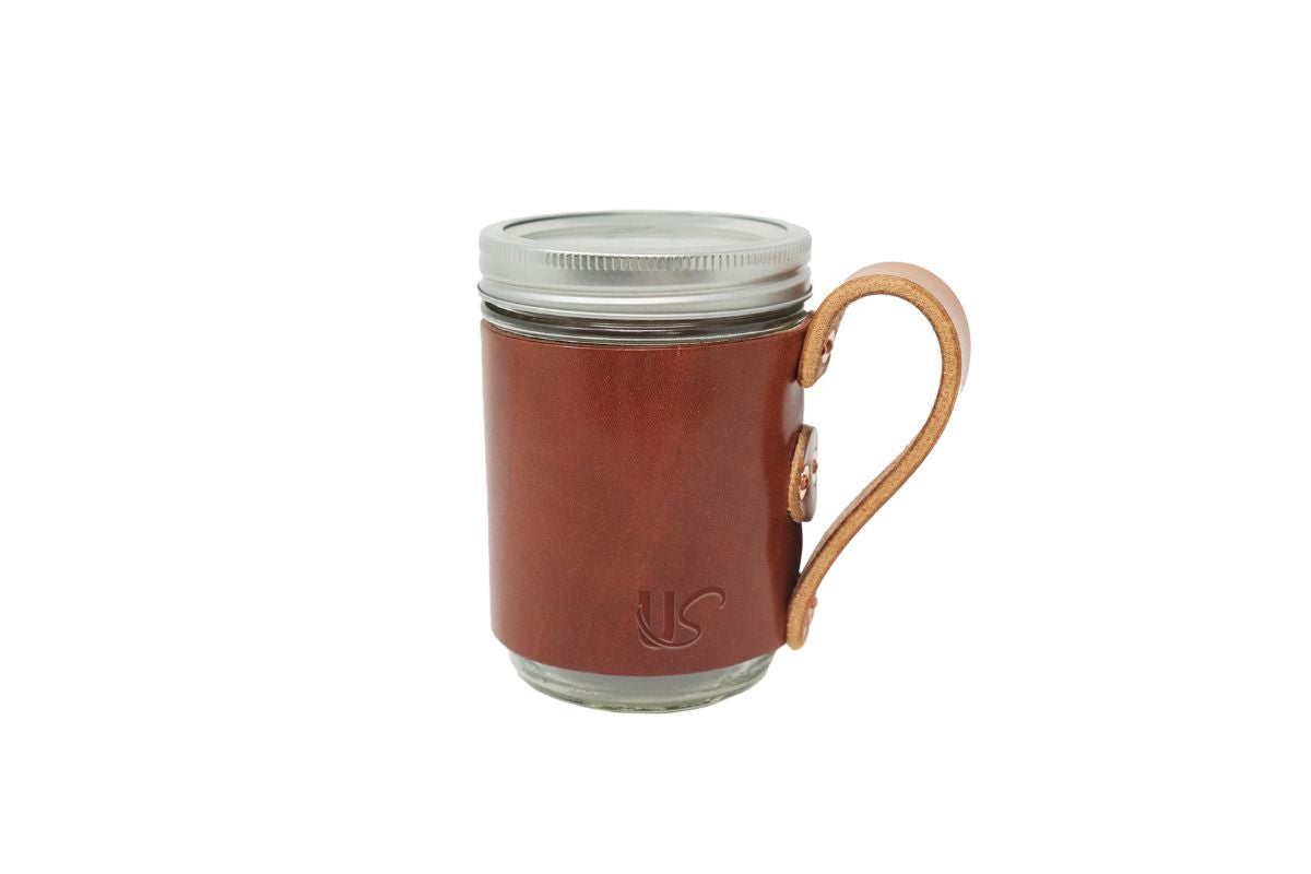 A glass mason jar mug wears a stylish brown leather sleeve with a handle.

