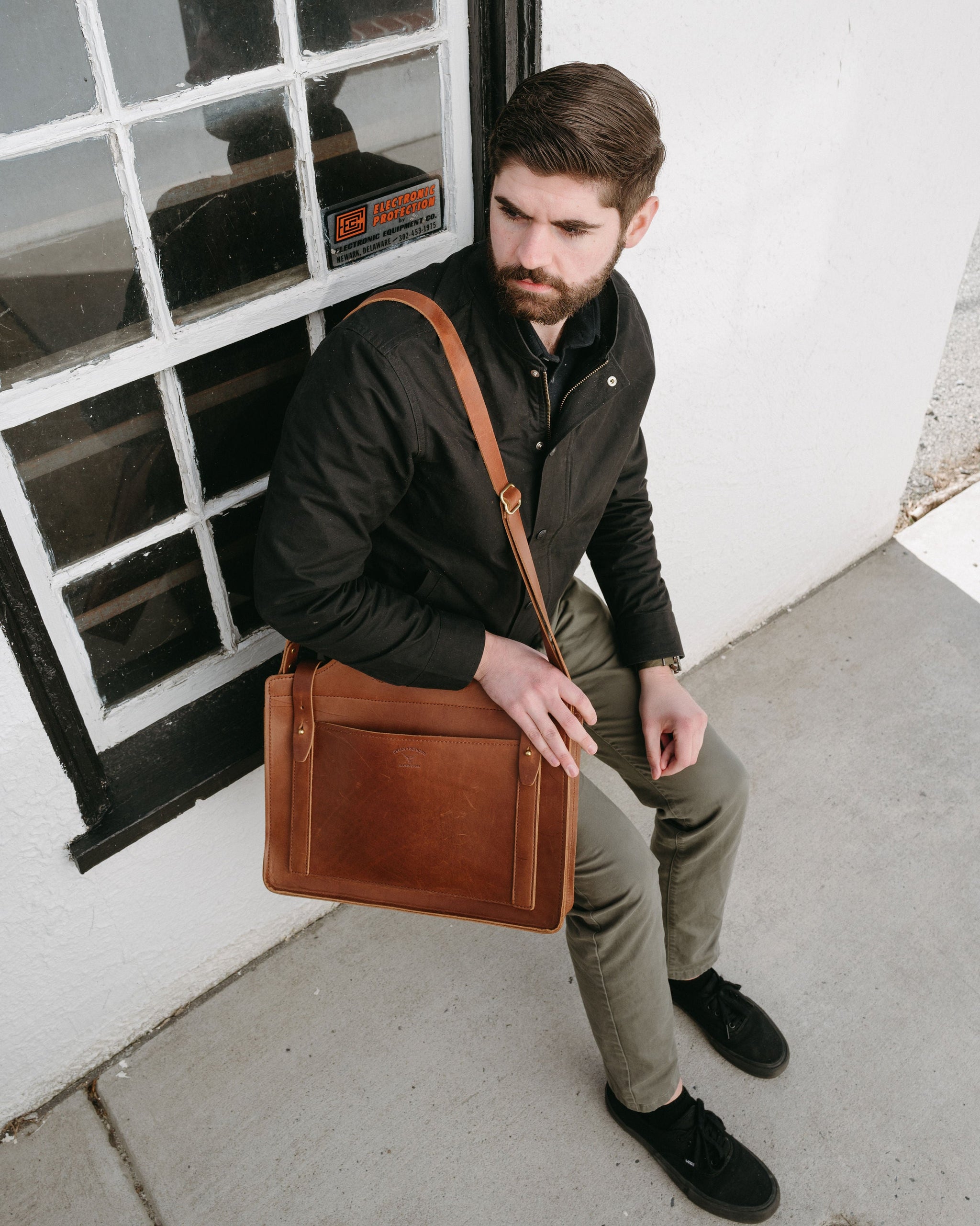 A bearded man models a brown leather messenger bag while sitting outside a white building with a window.
