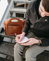 Man writes in a leather notebook with a gold pen, a stylish leather tote bag sits beside him on metal stairs.

