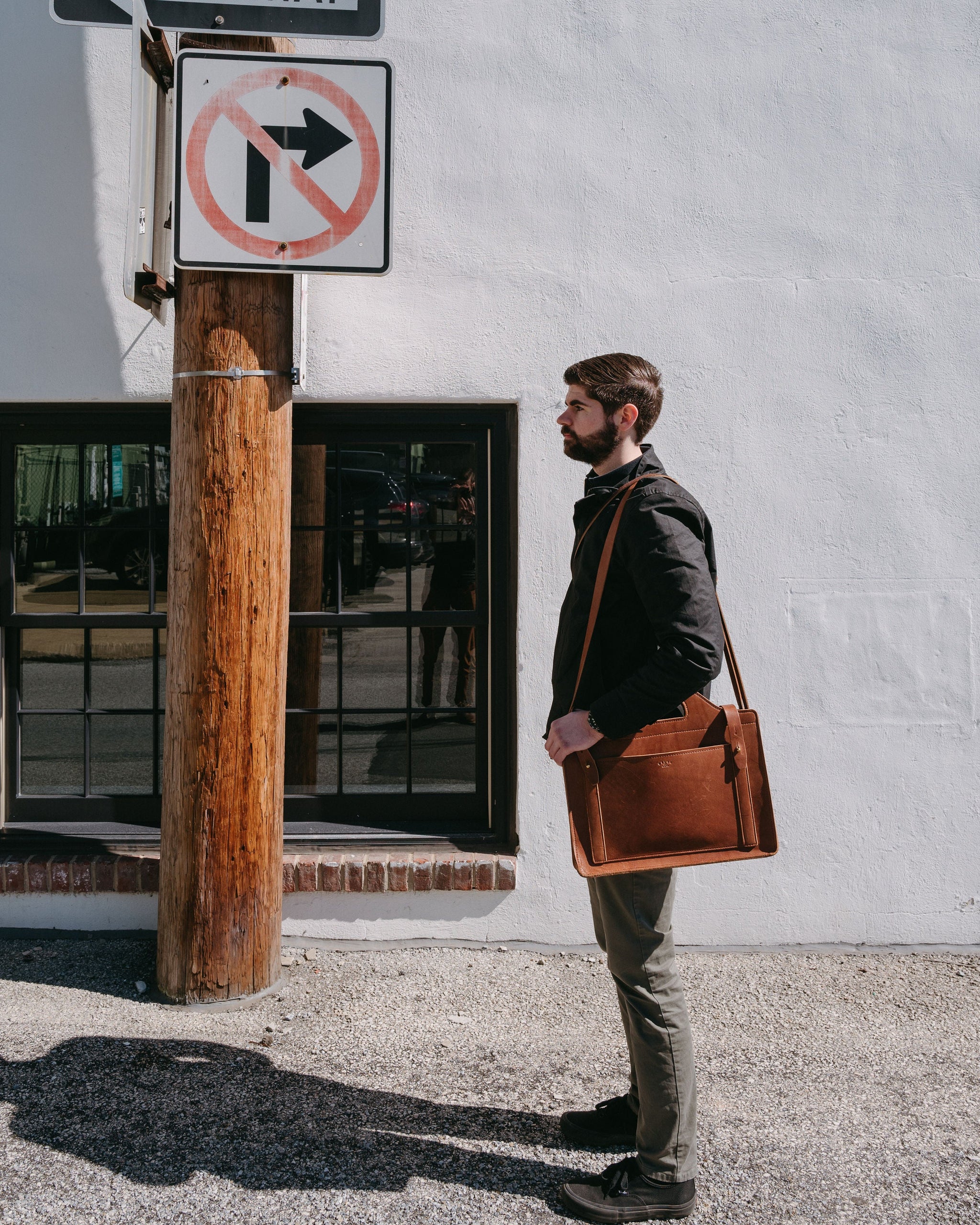A bearded man carries a leather messenger bag while standing next to a "no right turn" sign on a city street.
