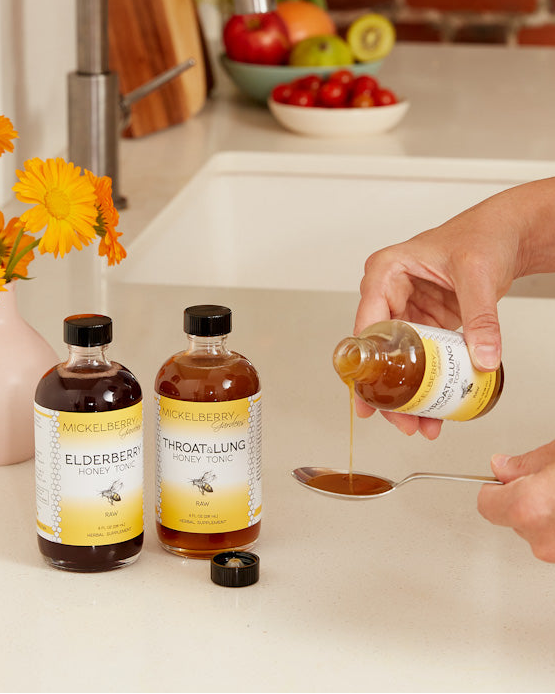 Hand pouring liquid from a bottle labeled 'Throat & Lung Honey Tonic' into a spoon on a kitchen counter with other bottles and fruits in the background.