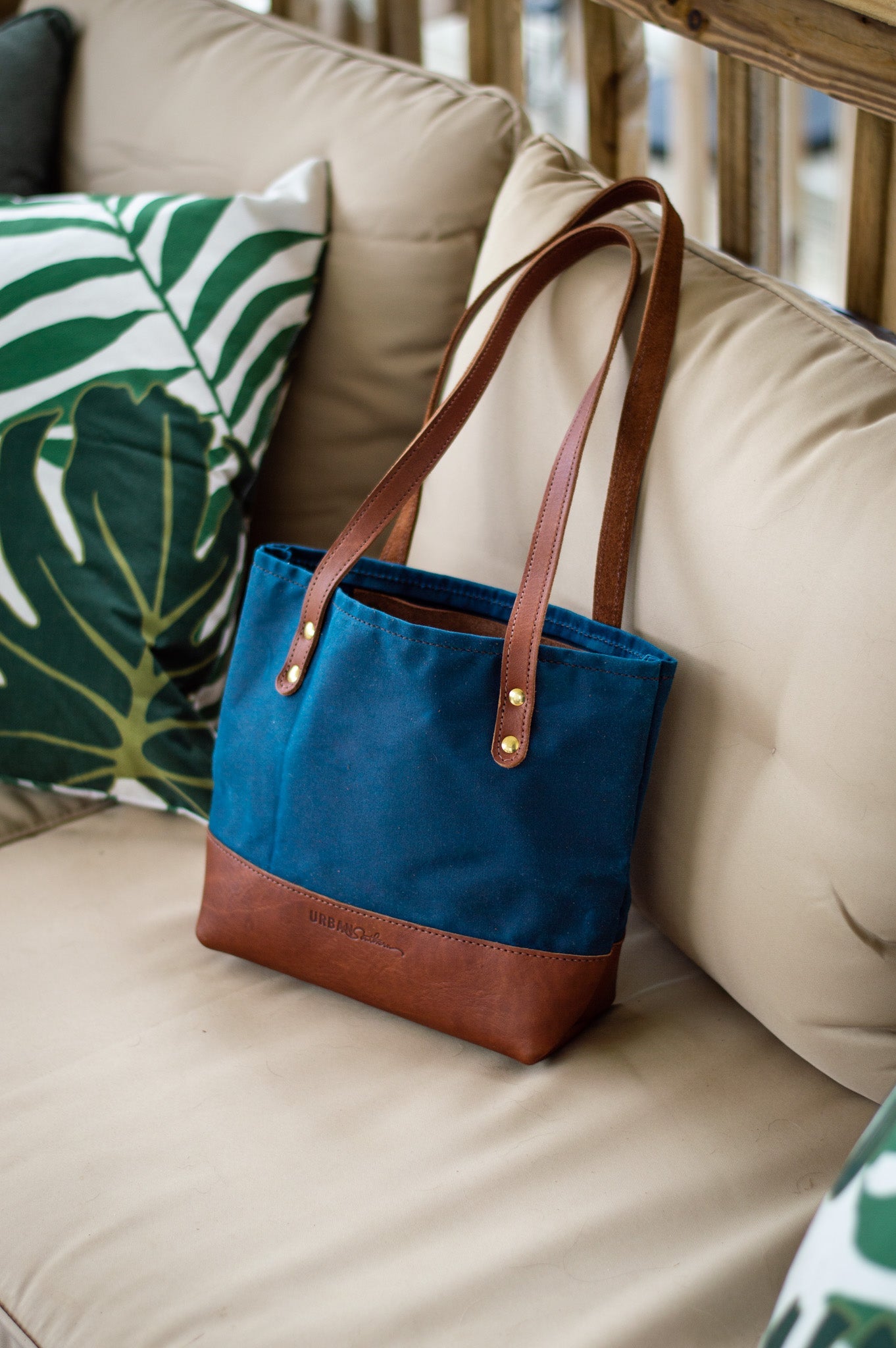 A blue canvas tote bag with leather handles and base sits on a beige outdoor couch next to a leaf-patterned pillow.
