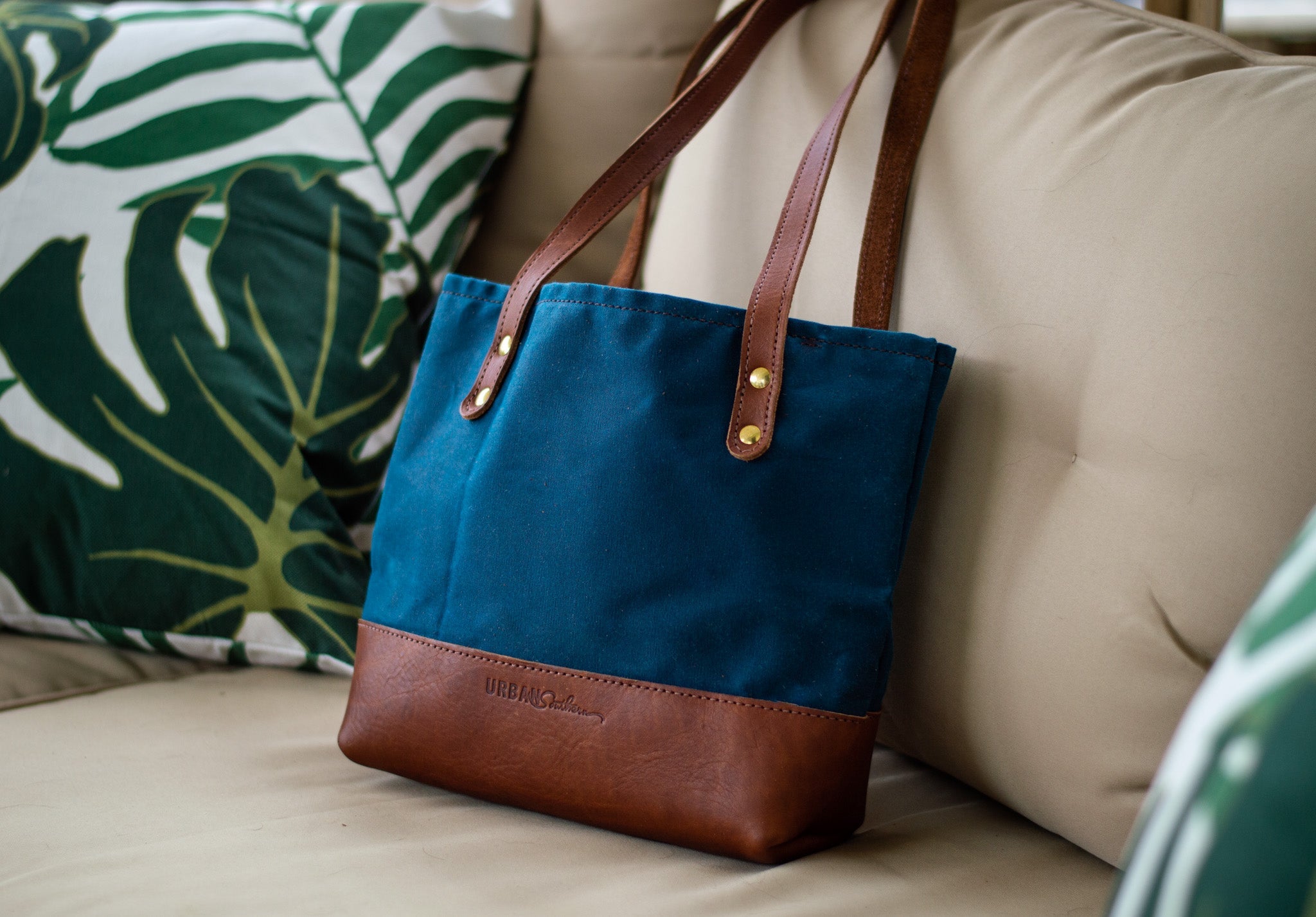 A blue canvas tote bag with brown leather accents rests stylishly on a beige sofa with a monstera leaf pillow.
