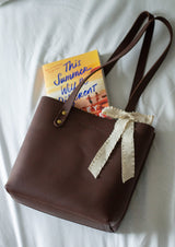 A brown leather tote bag with a ribbon detail holds the book "This Summer Will Be Different" on a white sheet.
