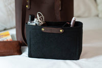 Organized travel enthusiasts use a black felt purse organizer insert from Urban Southern within a leather tote bag, holding essential items like hand sanitizer and earbuds.
