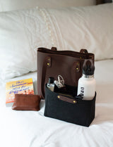 Organize your essentials with a leather tote bag, wallet, and organizer holding a water bottle, earphones, and sanitizer, styled on a white bed.
