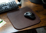 A Logitech wireless mouse rests on a brown leather mousepad next to a keyboard on a wooden desk.
