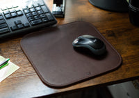 A Logitech wireless mouse rests on a brown leather mousepad next to a keyboard on a wooden desk.
