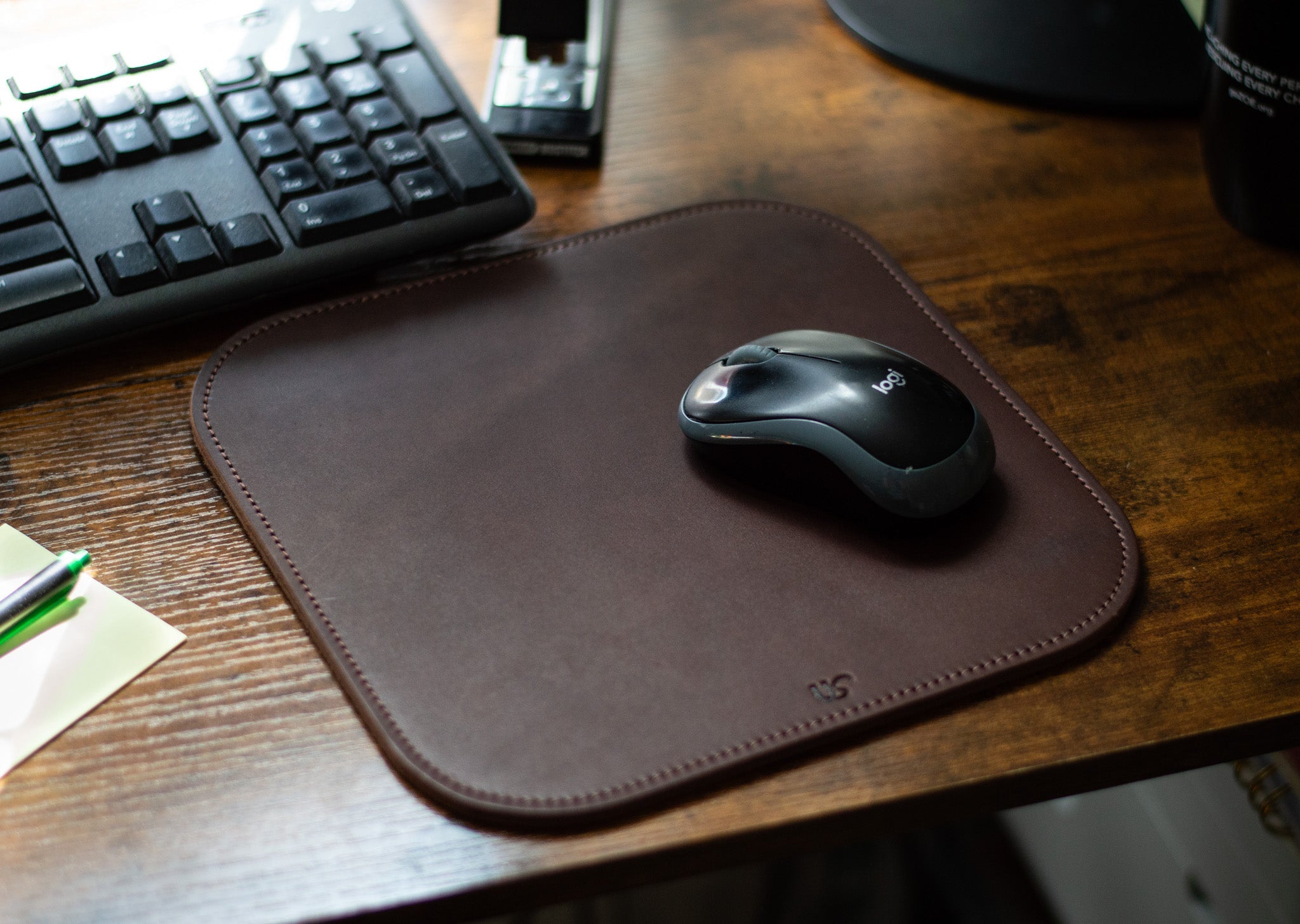 A Logitech wireless mouse rests on a brown leather mousepad next to a keyboard on a wooden desk.
