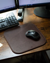 A Logitech mouse rests on a stylish brown leather mousepad beside a computer keyboard and stapler on a wooden desk.
