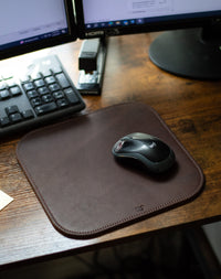 A Logitech mouse rests on a stylish brown leather mousepad beside a computer keyboard and stapler on a wooden desk.
