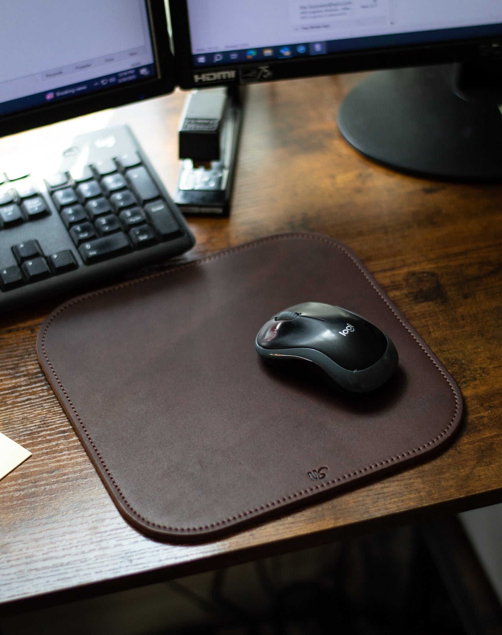 A Logitech mouse rests on a stylish brown leather mousepad beside a computer keyboard and stapler on a wooden desk.
