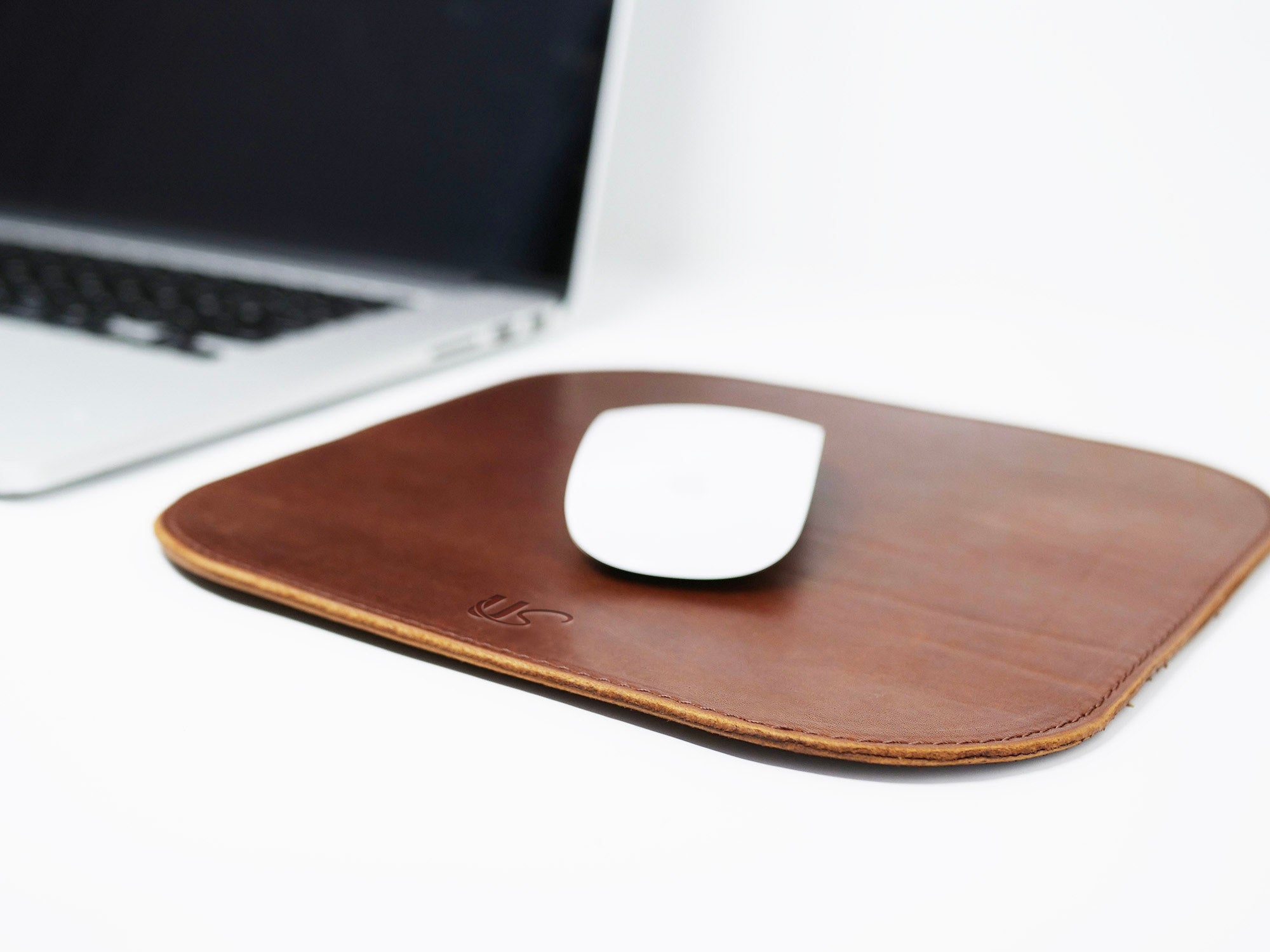 A white wireless mouse rests on a handcrafted brown leather mousepad next to a blurred laptop.
